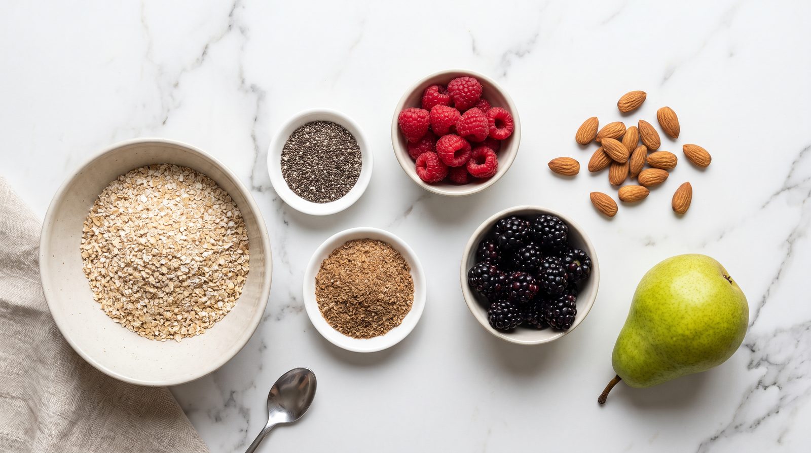 High fiber breakfast ingredients on a marble surface: chia seeds, oat bran, raspberries, blackberries, ground flaxseed, almonds and a pear