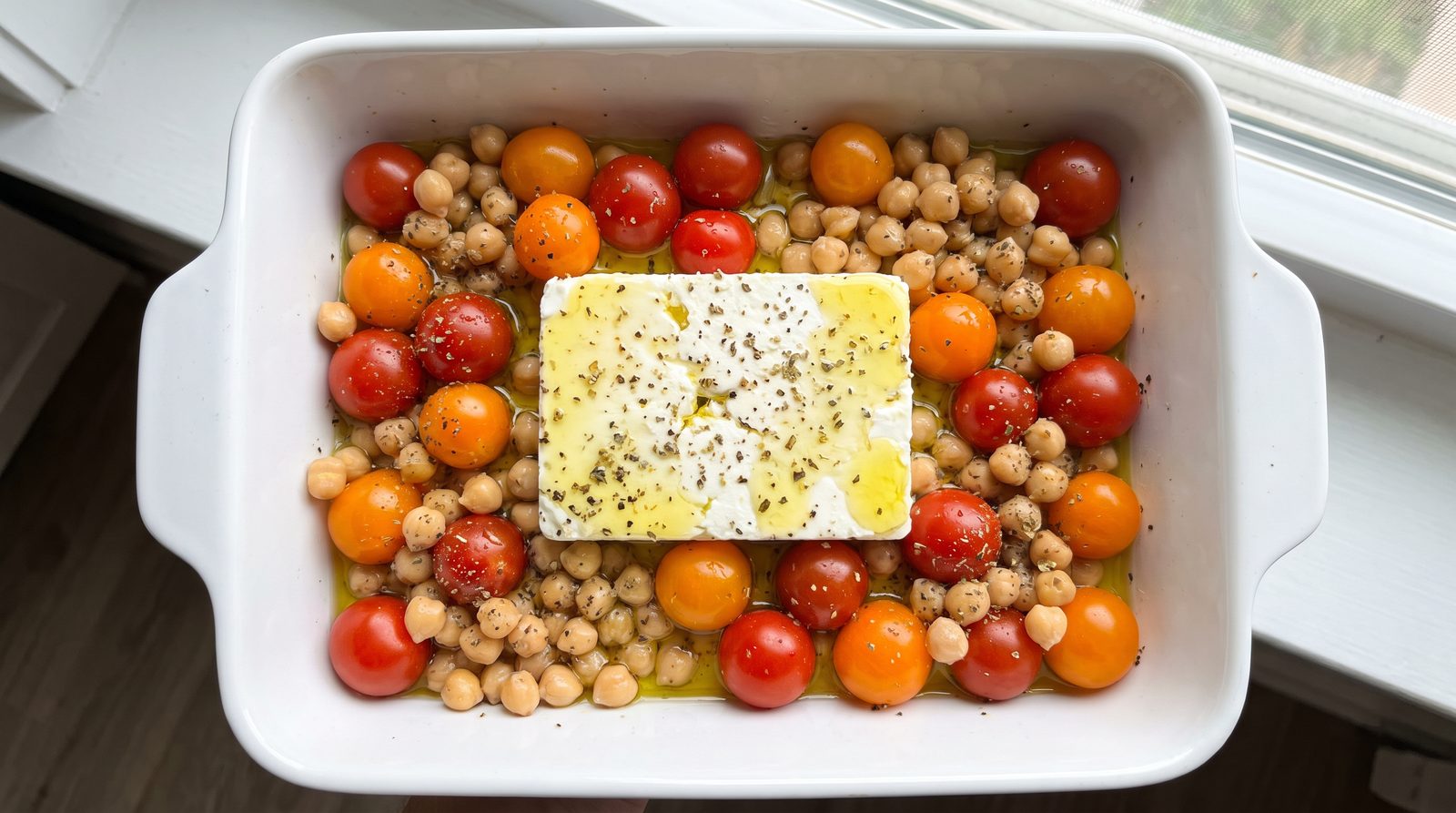 A baking dish before roasting with a block of feta surrounded by cherry tomatoes and chickpeas in olive oil