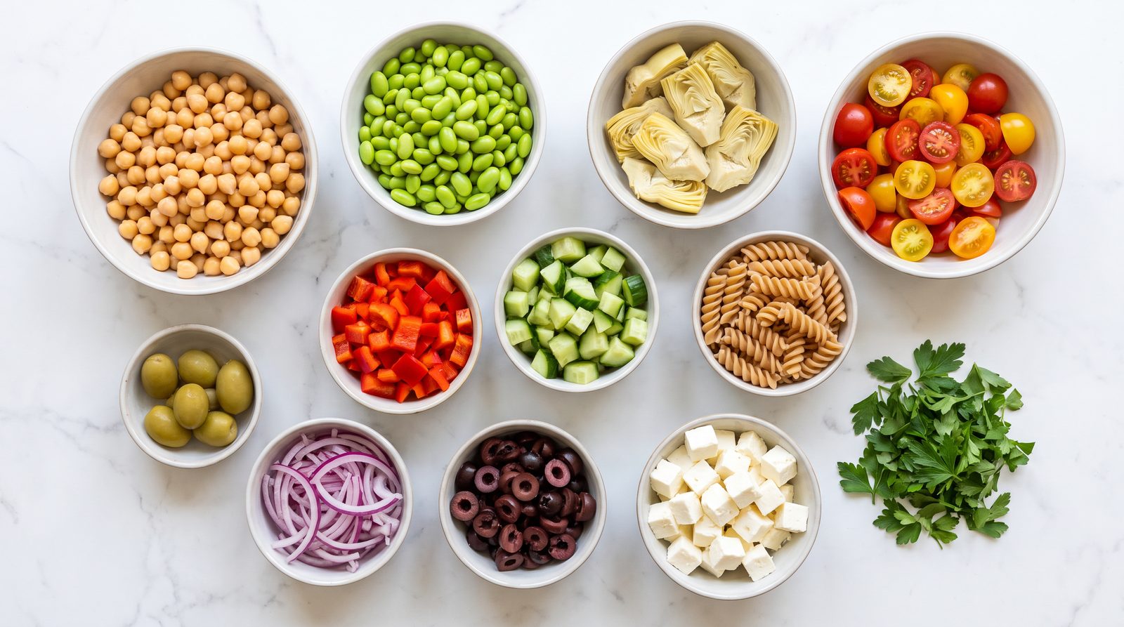 Overhead view of prepped pasta salad components in separate bowls — chickpeas, shelled edamame, artichoke hearts, cherry tomatoes, red pepper, cucumber, red onion, olives, feta and parsley, with a smaller bowl of whole wheat fusilli