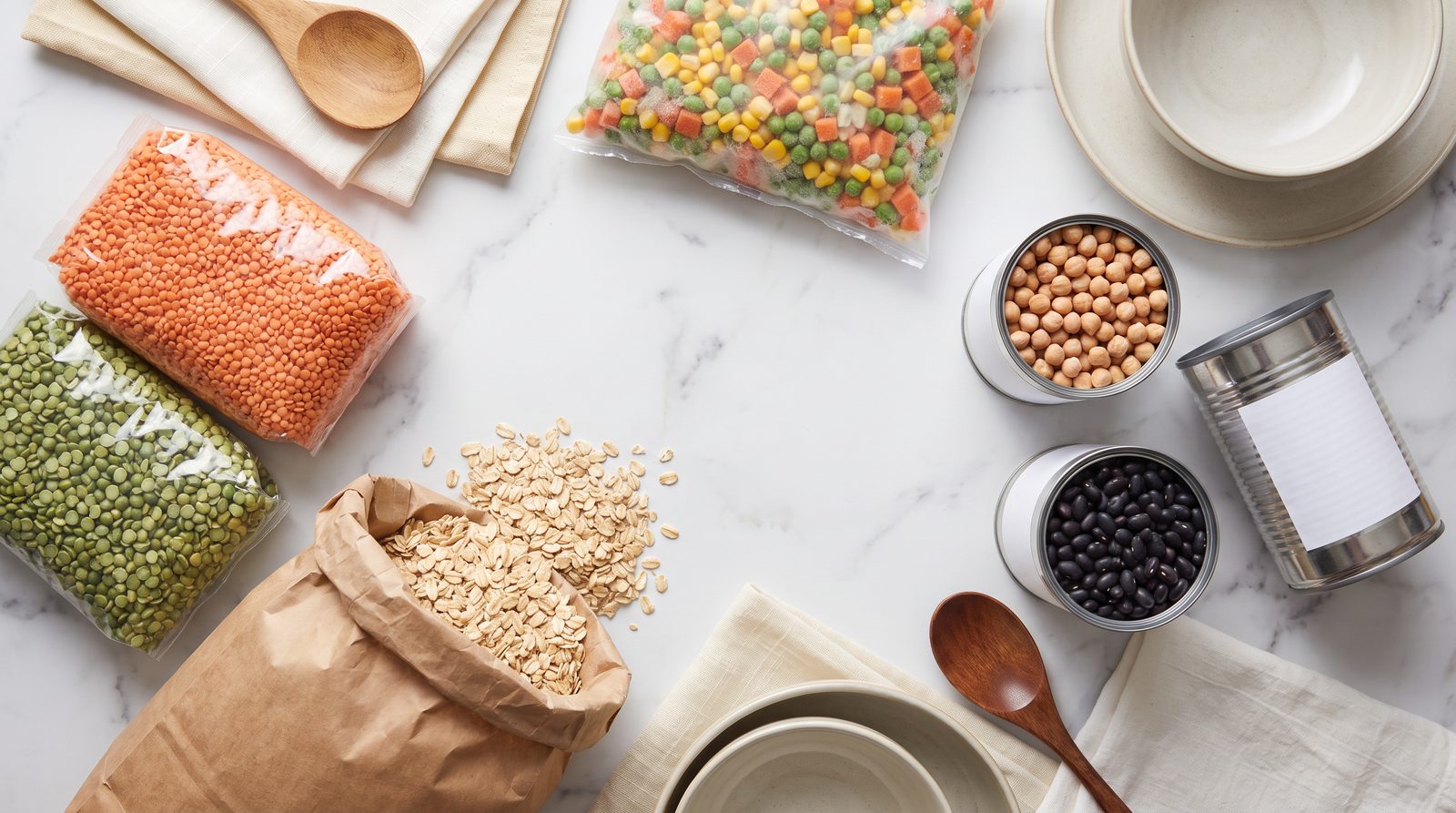 Cheap high fiber staples on white marble: dried red lentils, split peas, rolled oats, tinned chickpeas, tinned black beans and frozen vegetables