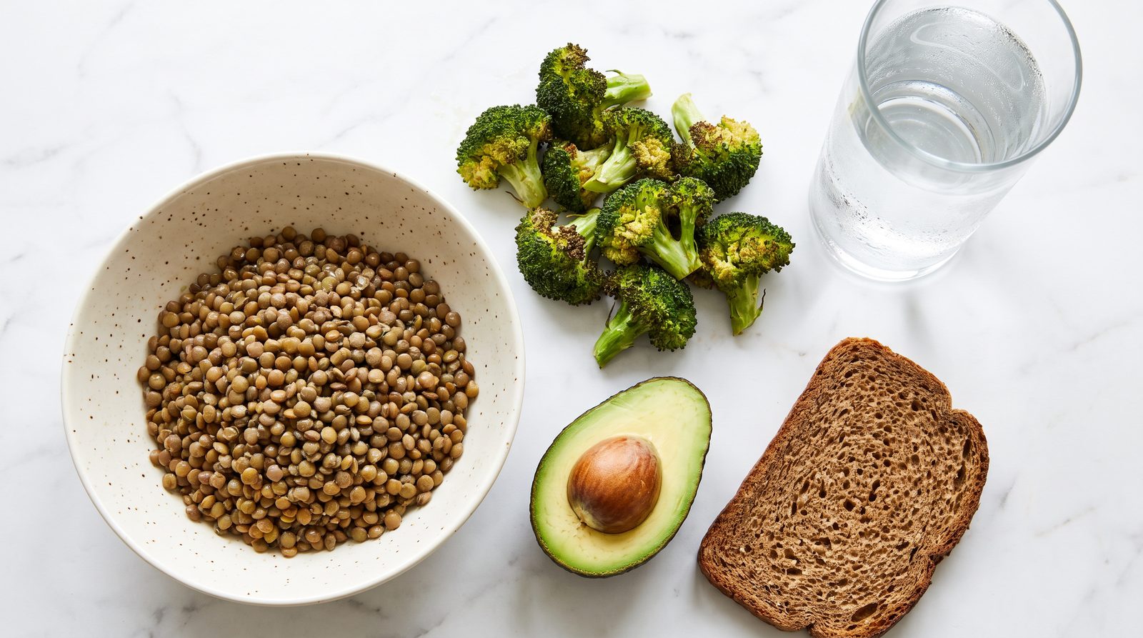 A simple high fiber meal on white marble: a bowl of lentils, roasted broccoli, half an avocado, dark wholemeal bread and a glass of water