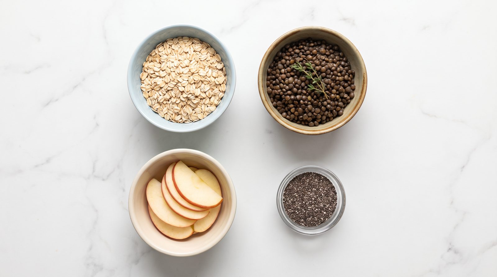 Four different fiber types in separate bowls on white marble: rolled oats, cooked lentils, sliced apple with skin, and chia seeds