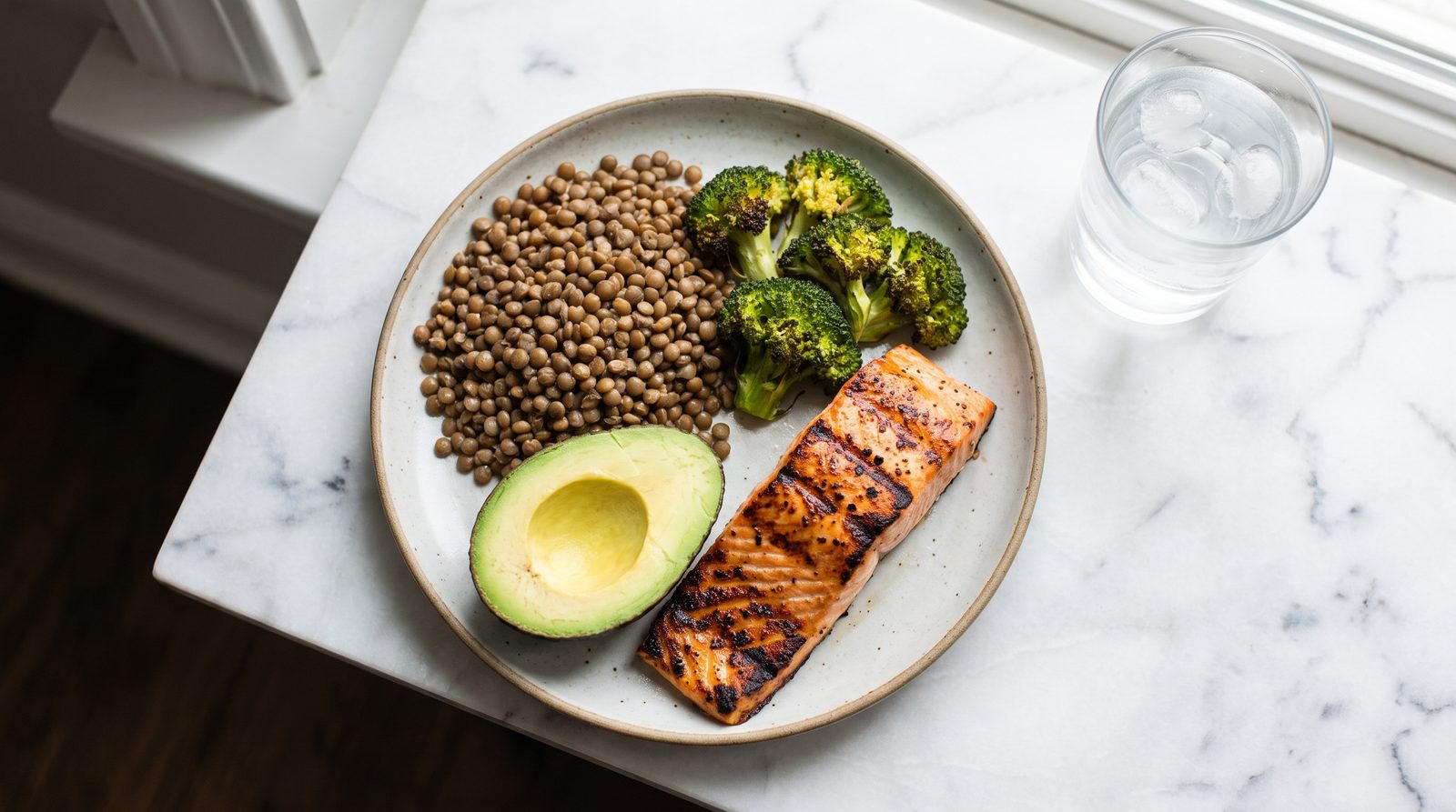 A GLP-1 friendly plate on white marble: grilled salmon, cooked lentils, roasted broccoli and half an avocado with a glass of water